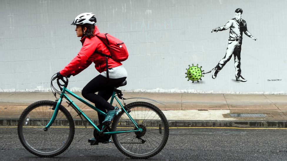 Woman cycling by graffiti showing a man chained to a coronavirus