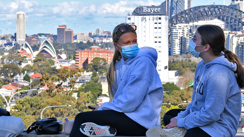 Two girls wearing face masks sit above the Bradfield Highway in the suburb of North Sydney with the city in the background on July 07, 2021 in Sydney, Australia.