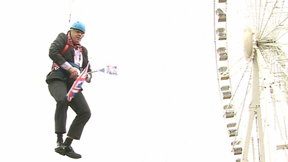 Boris Johnson dangles from a zip wire in front of a big wheel, while holding union jack flags and wearing a suit